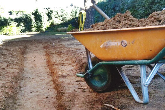 Una carretilla de obra de acero pintado cargada de tierra junto a una zanja de drenaje.